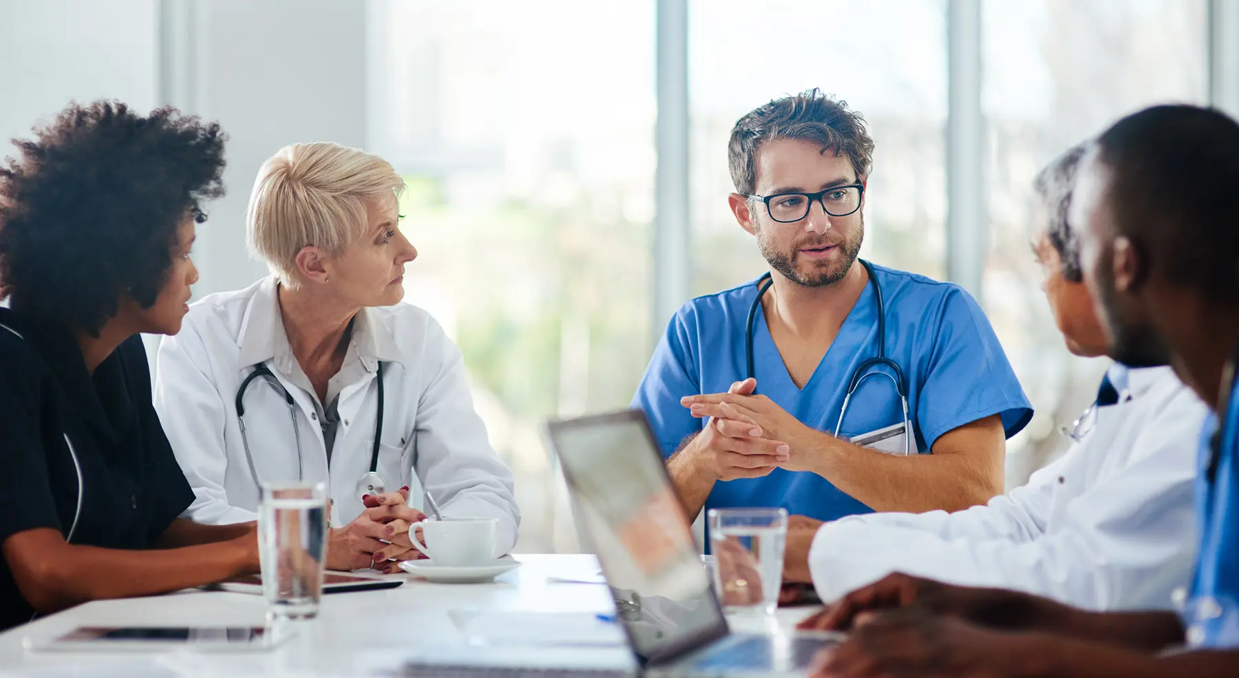 A medical professional wearing scrubs talks to their colleagues around a table