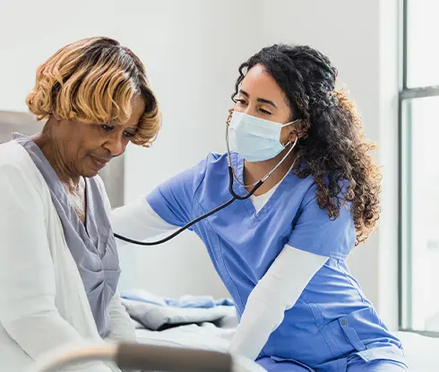 A woman wearing scrubs and a facemask uses a stethoscope on an elderly patient